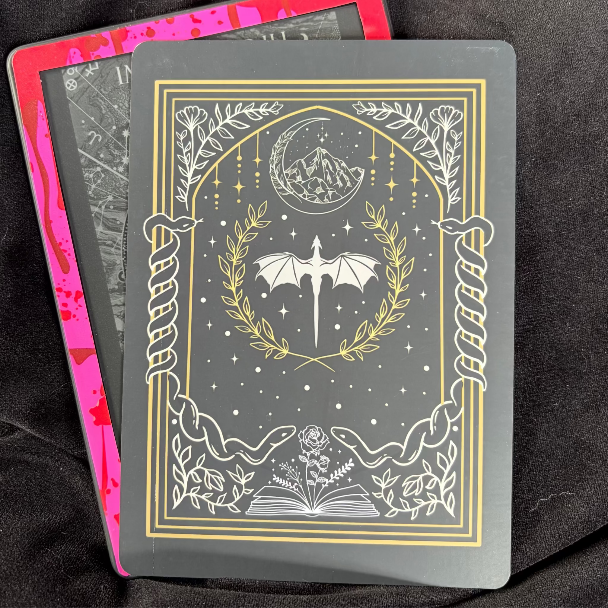 Black romantasy Kindle backing insert with gold and white illustrations of a dragon, winged dagger, moon, flowers and open book
