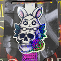 Glitter magnetic bookmark featuring a stitched white bunny with X-shaped eyes sitting on a skull surrounded by leaves and a rose
