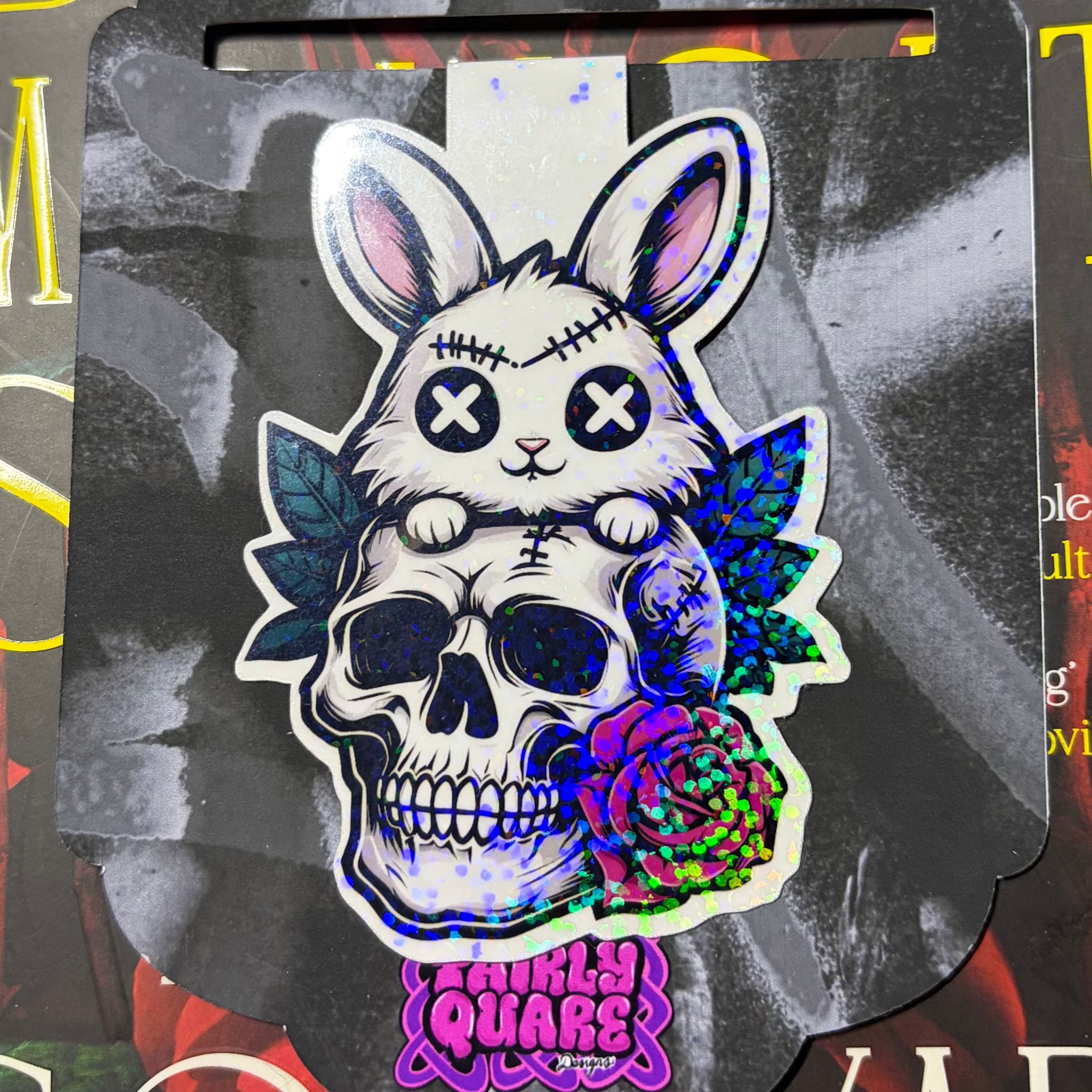 Glitter magnetic bookmark featuring a stitched white bunny with X-shaped eyes sitting on a skull surrounded by leaves and a rose
