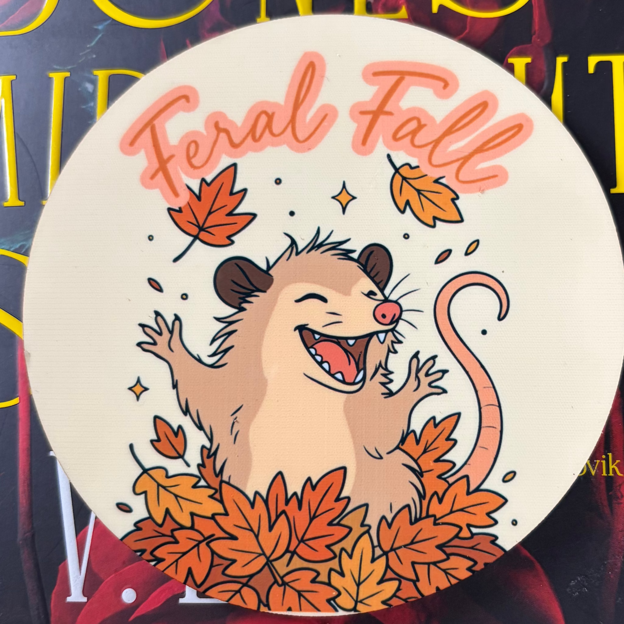 Round cream Feral Fall coaster featuring an excited cartoon opossum surrounded by orange autumn leaves
