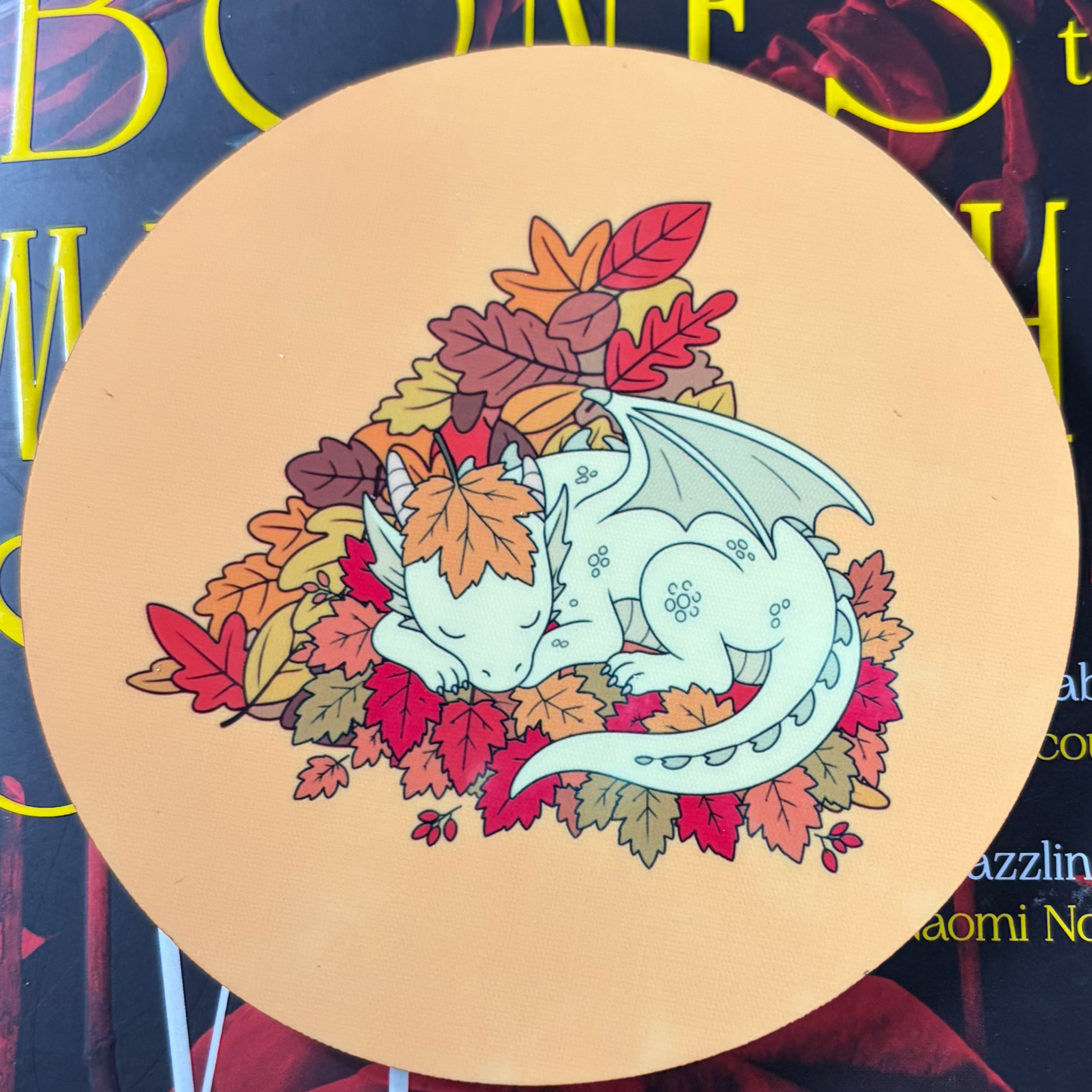 Round orange coaster featuring a cream-coloured sleeping dragon curled up in a pile of colourful autumn leaves
