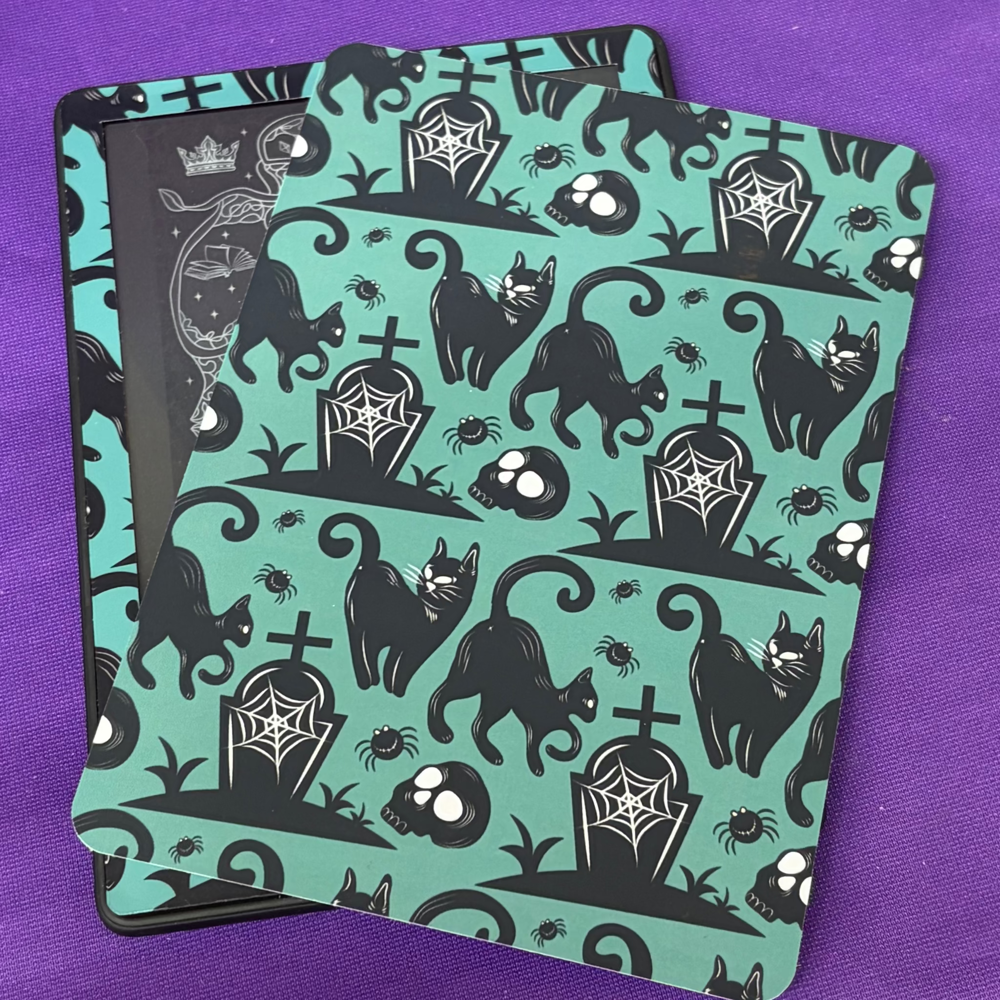 Teal and black Kindle backing insert patterned with spooky cats, skulls, gravestones, spiders and cobwebs
