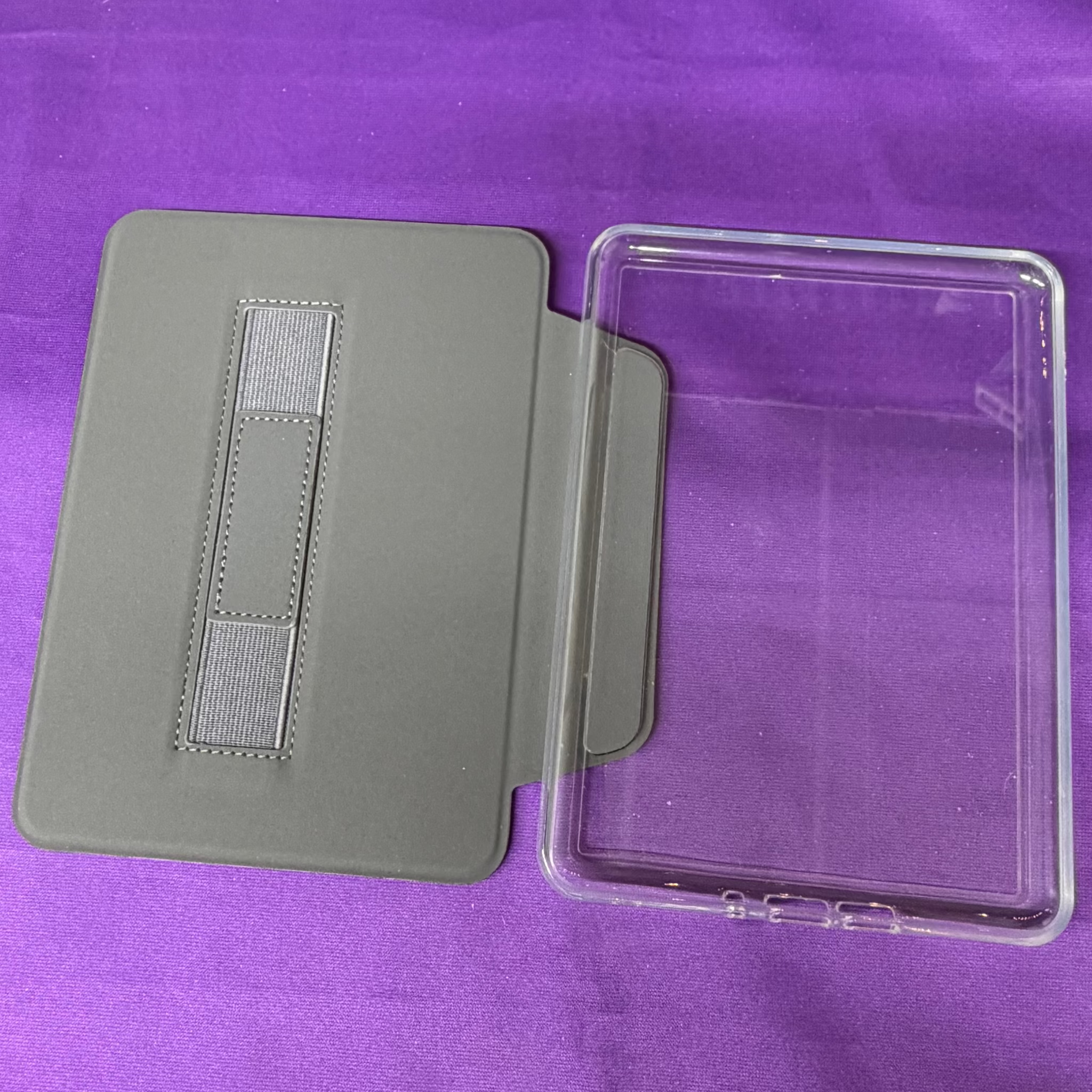 A clear protective tablet case with a built-in gray kickstand, shown open on a purple fabric background.