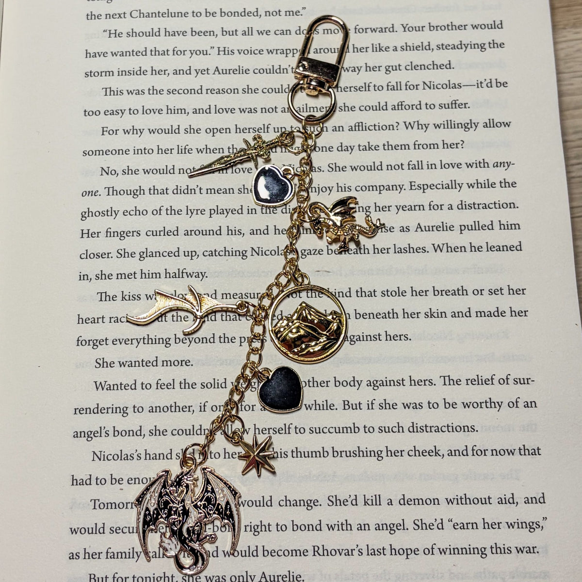 Dragon Rider: Gold Kindle charm with black enamel dragon, dagger, mountain medallion, dragon wing, black hearts and star charms on a swivel clasp, laid on an open book