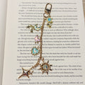 Starlit Daydream: Gold Kindle charm with iridescent blue moon, pink butterfly, aqua heart, shooting star and holographic sun charms on a swivel clasp, laid on an open book