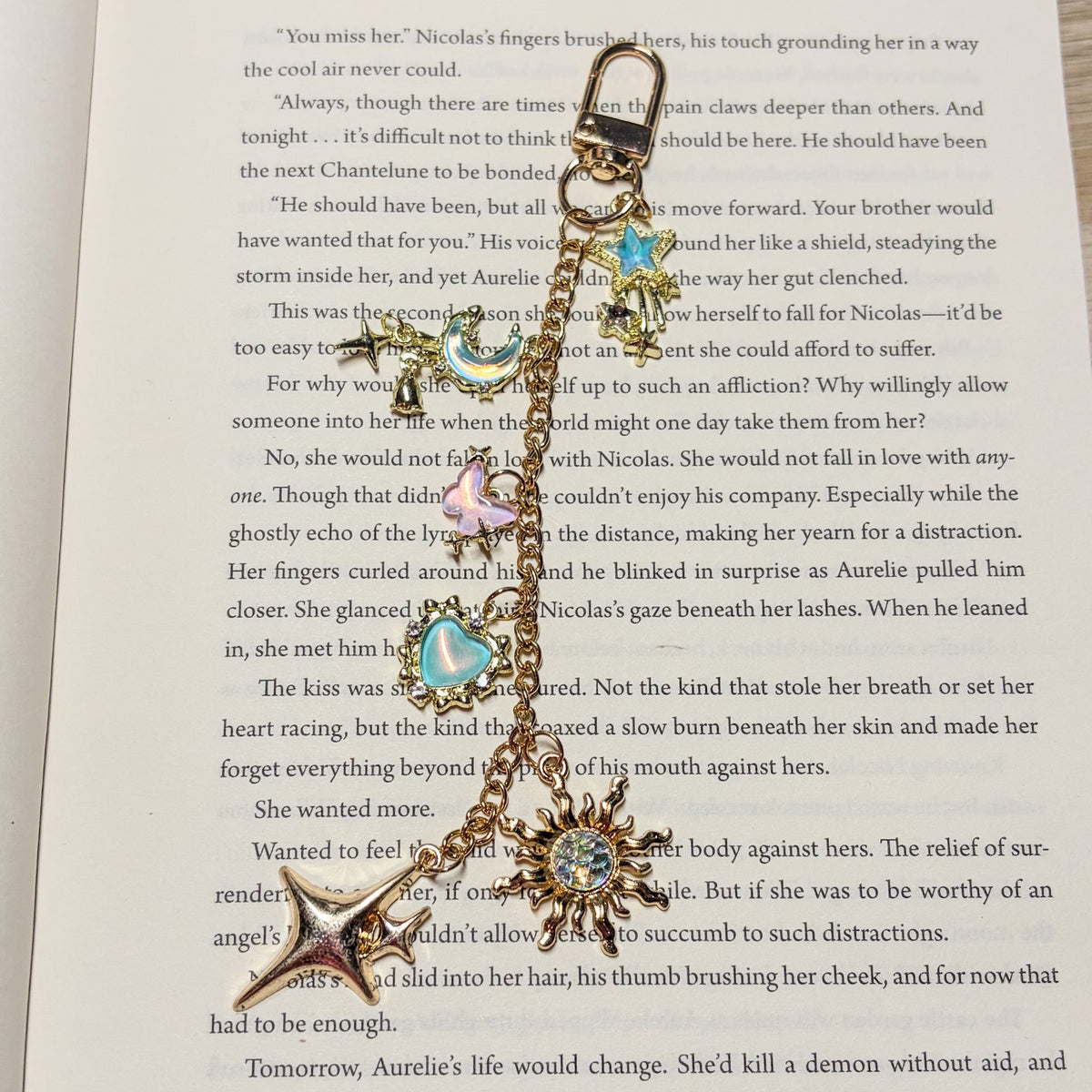 Starlit Daydream: Gold Kindle charm with iridescent blue moon, pink butterfly, aqua heart, shooting star and holographic sun charms on a swivel clasp, laid on an open book